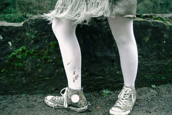 Young woman wearing skirt with feathered frontpiece, white tights soiled from muddy path and camouflage trainers stands side on