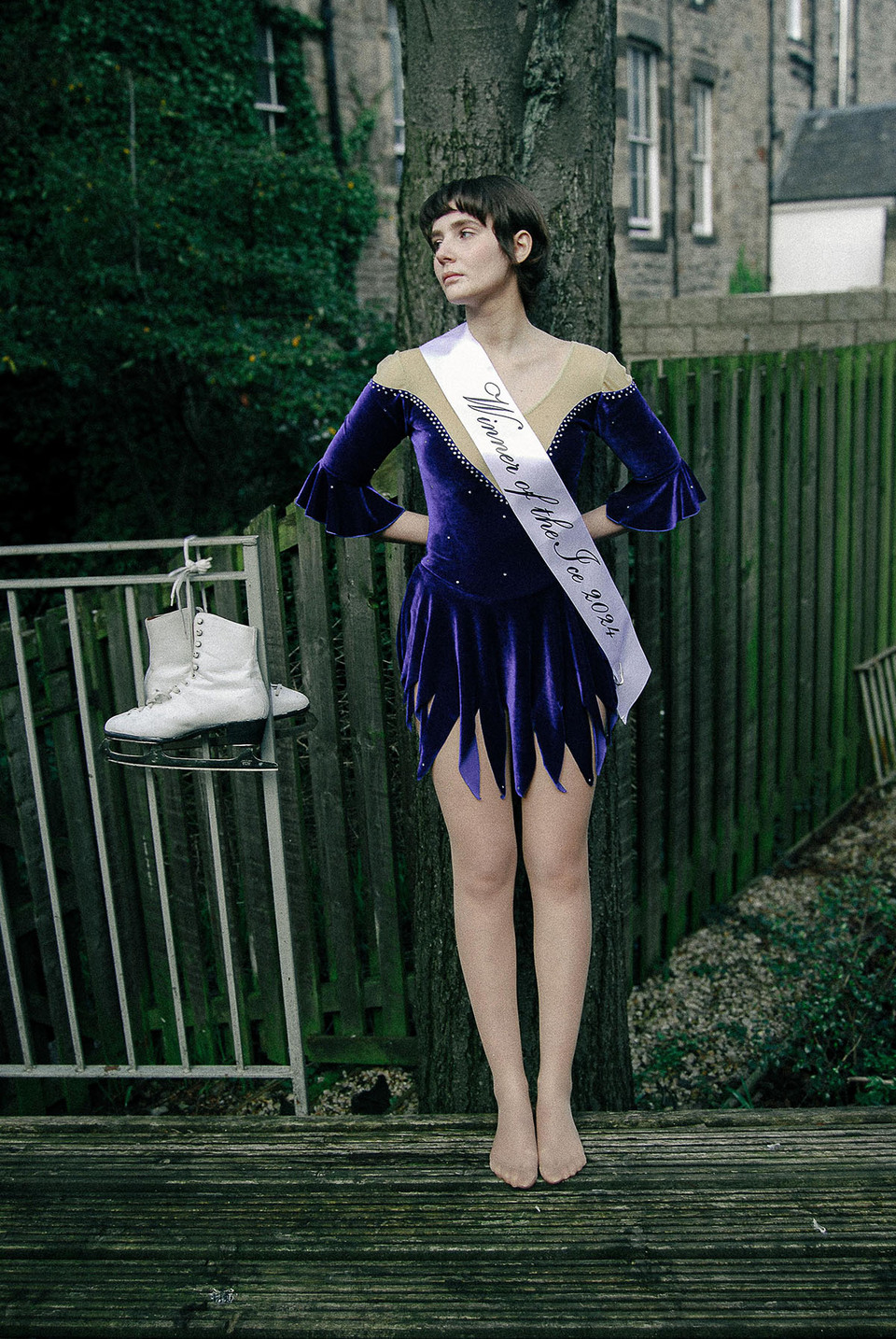 Young woman looks to her right wearing an ice skaters deep blue dress with 'Winner of the Ice 2024' sash leans against a tree while her ice skating boots hang from a railing beside her