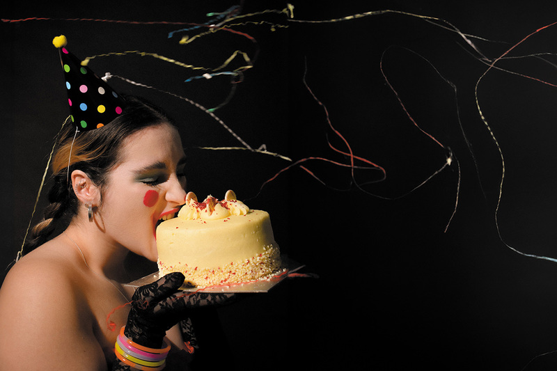 Young woman wearing lace gloves, coloured bangles, party hat and strapless dress bites into a whole white chocolate cake she holds before her face while party streamers fly through the air