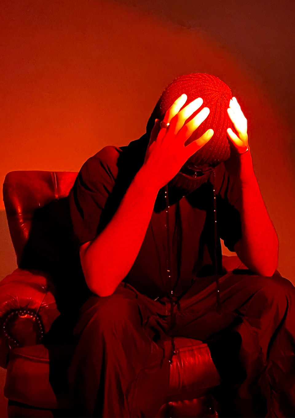 A man seated, holding his balaclava-clad head in his hands is bathed in a red light