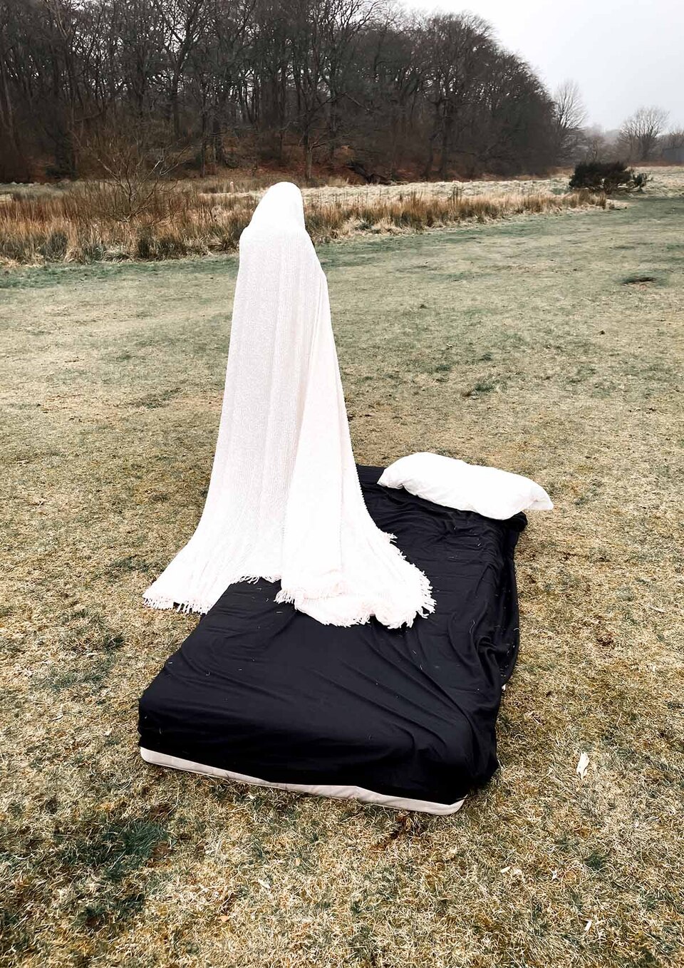 Rear view of a human figure wrapped from head to foot in a white blanket steps off a black-sheeted mattress in an open field