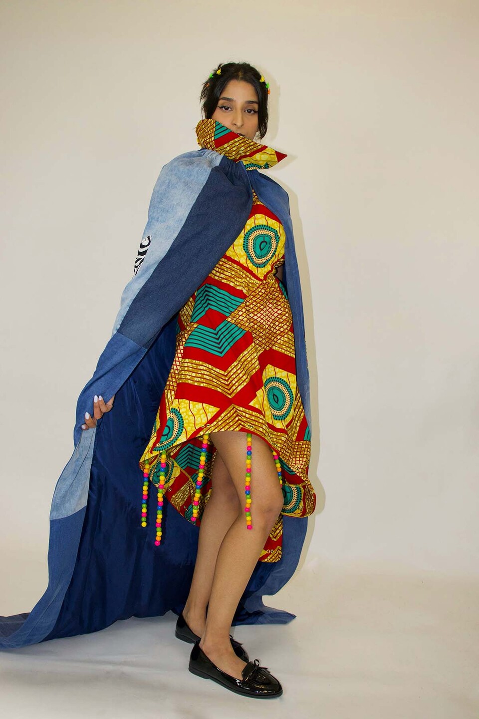 Woman models an African print dress in yellow, red, brown and teal with strings of coloured beads hanging from the hem and a large floor length upcycled denim cape.