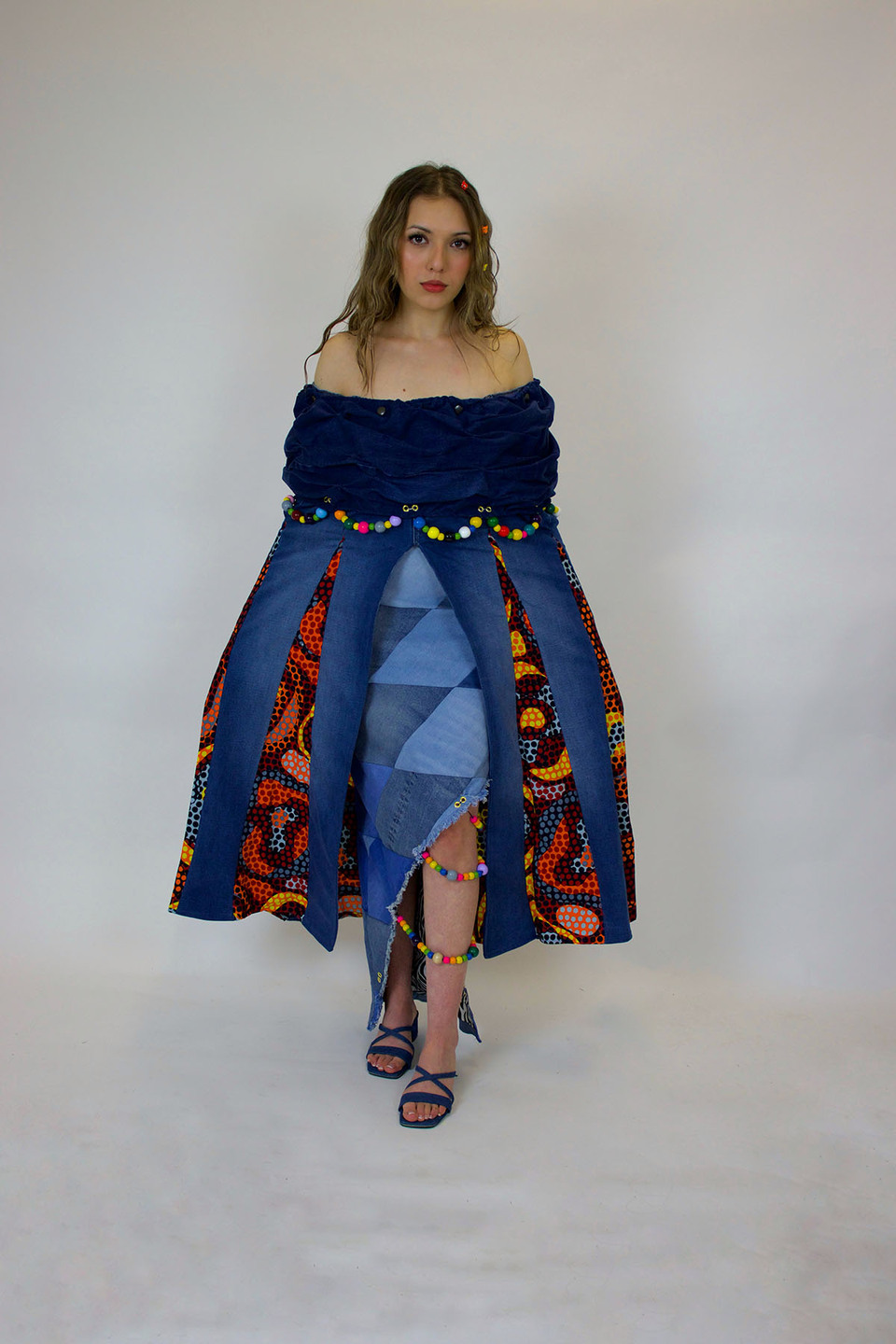 Woman models an off the shoulder African print pleated dress in blue, red, orange and pink with upcycled denim pleats and coloured beading around the waist.