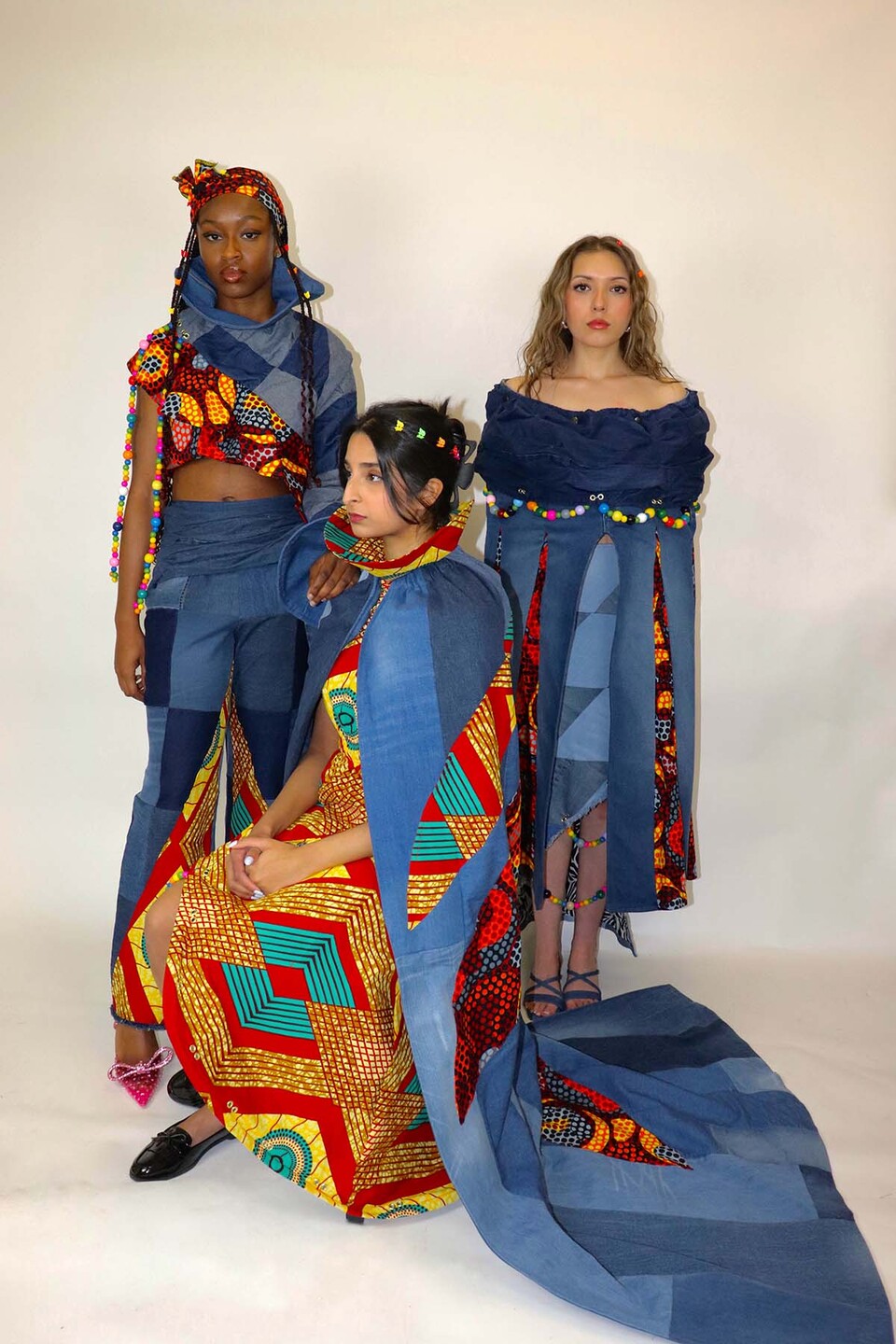 Three women model two African print dresses, and top and trousers in blue, red, brown, orange and teal with upcycled denim sections and beading from 'The Southside' by Chrystal Dube.