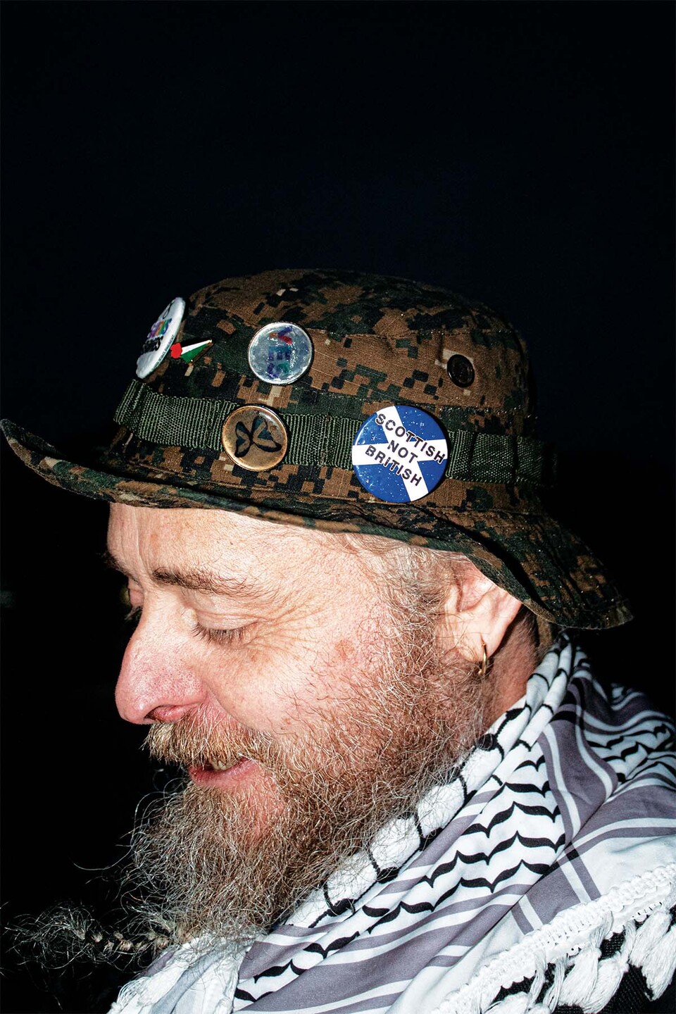 Old bearded man wearing a keffiyeh and a hat with prominent 'Scottish not British' badge attached