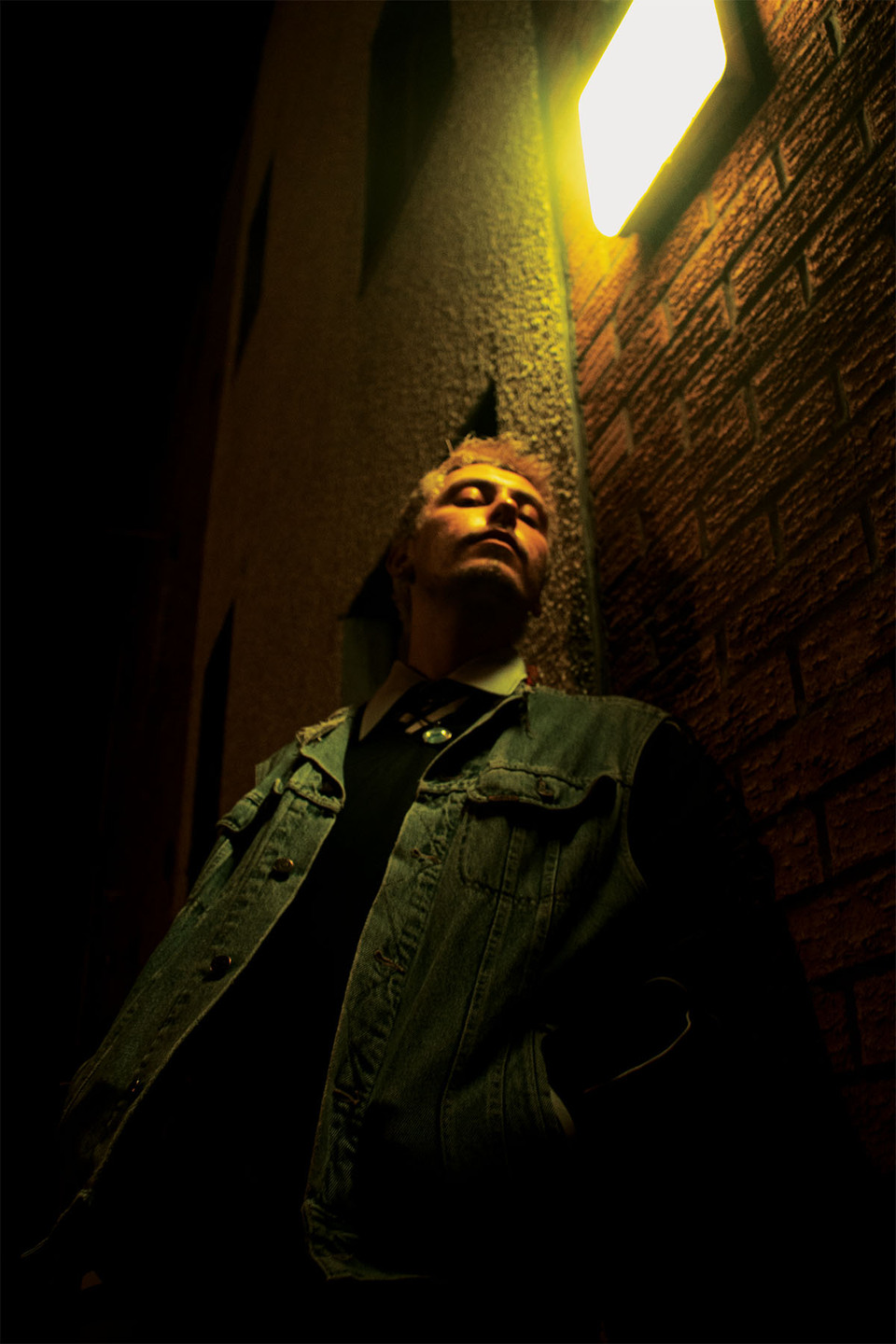 Man in a denim jacket stands under an external wall light looking down
