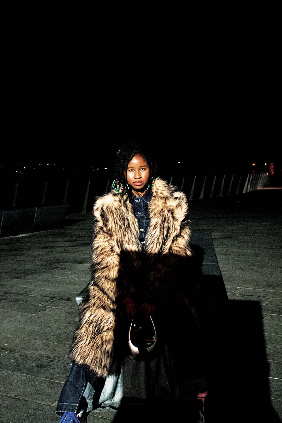 Young woman wearing denim jeans and shirt under a long fur coat holds a handbag in front of her at night on a riverside walkway