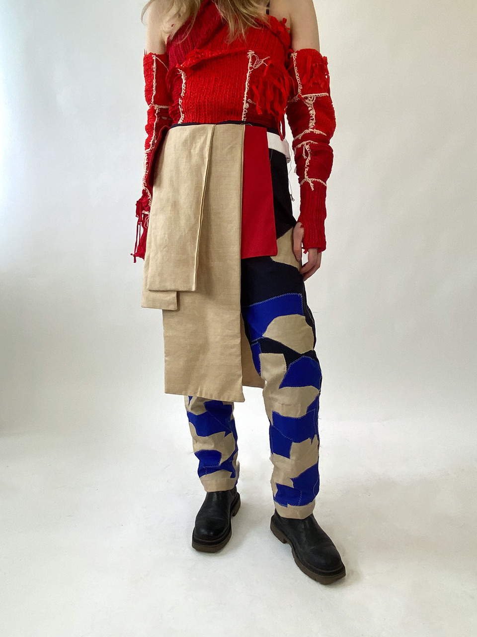 Gender neutral clothing design featuring patchwork straigh leg trouser in black, blue and khaki, red shoulderless woolen jumper with exposed white stitched seams and belt slung draped fabric 