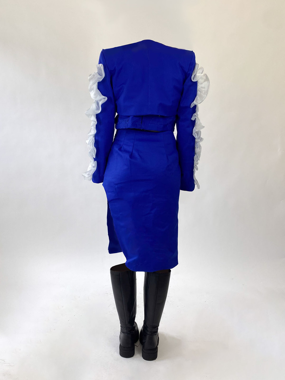 Rear view of blue collarless skirt suit with crimped white lace details on the outer sleeves and knee length black flat soled boots by Gosia Czerwinska