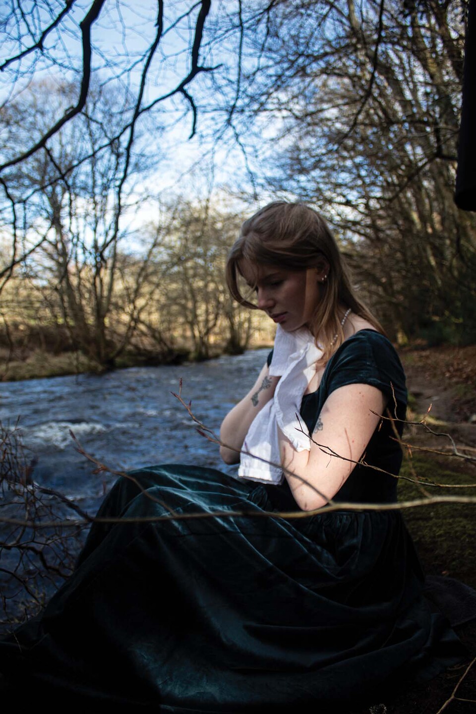 Young woman in long dark green dress sits beside a river holding a white shirt to her chest 