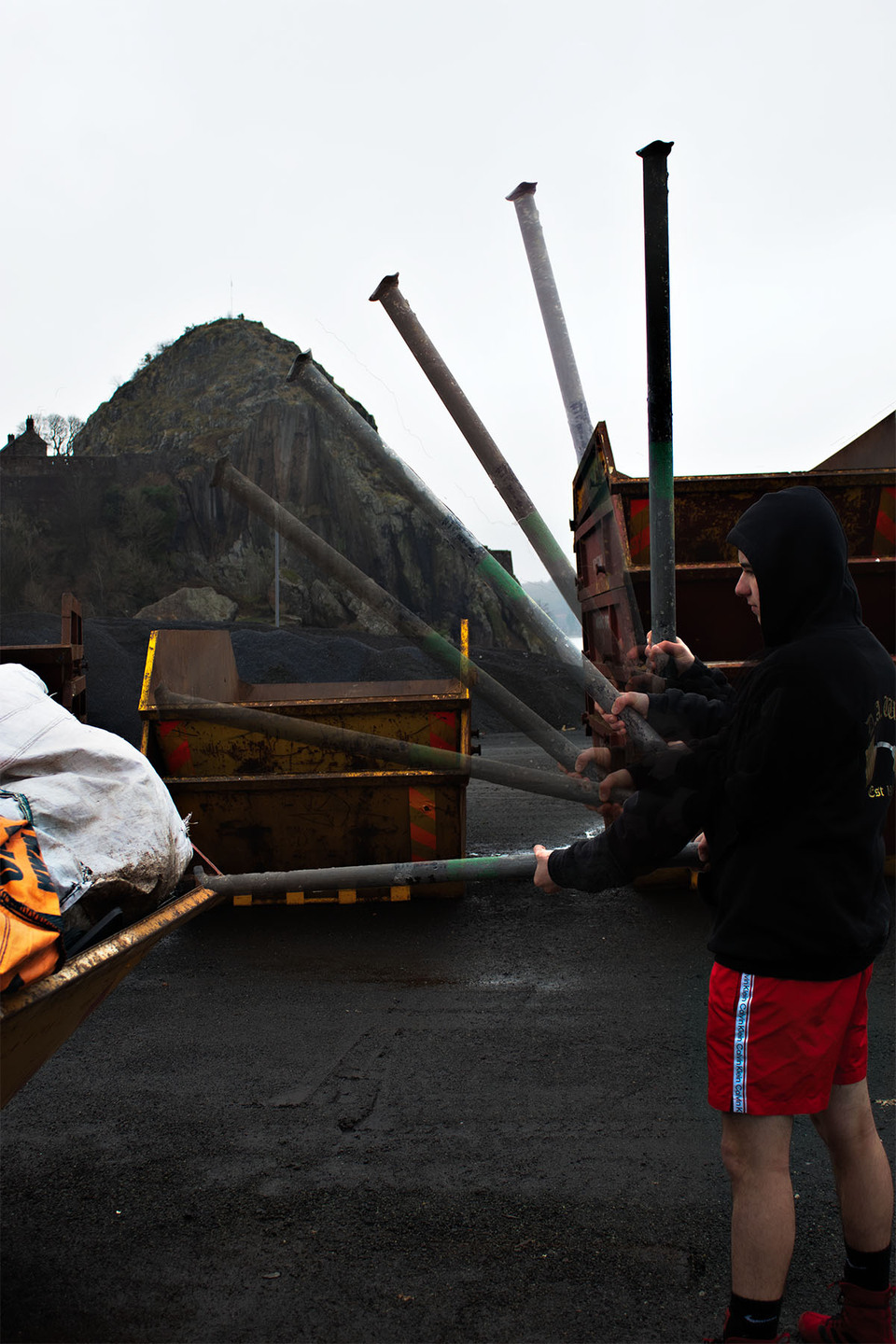 Young man in black hoodie and red shorts swings a metal pole in an industrial site 