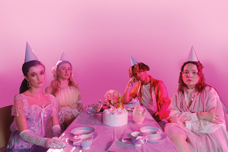 Three women and one man wearing party hats sit at a table bathed in pink light with sombre expressions