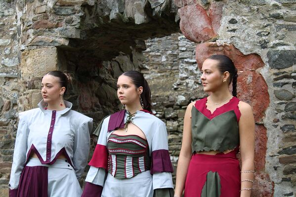 Three women model Karen Heath designs - dress, skirts, trousers, top and jacket standing before the gateway to a ruined castle