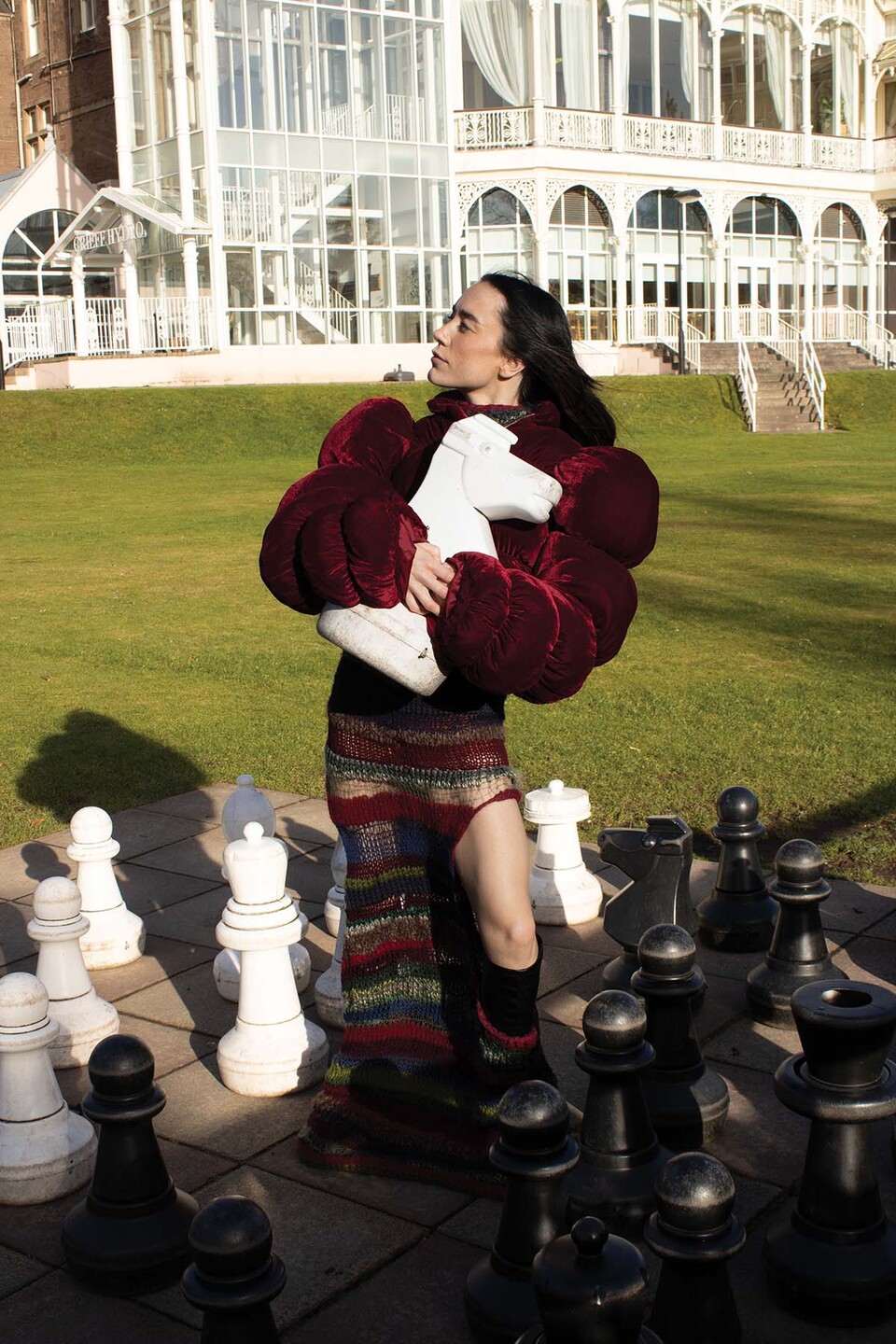 Woman in padded red jacket and knitted maxi skirt slit on one side stands on an outdoor giant chessboard, holding the white knight to her chest