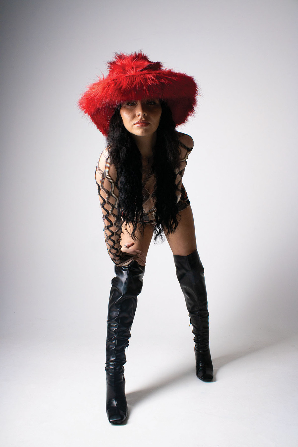 Woman leans forward in over the knee leather boots, large red fur hat, leather underwear and curving lattice-like top
