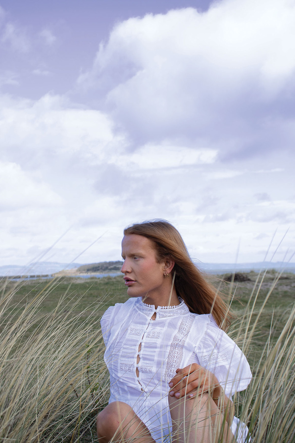Model wears a white collarless dress with lace panels in neck and front sitting in long grass looking out to sea