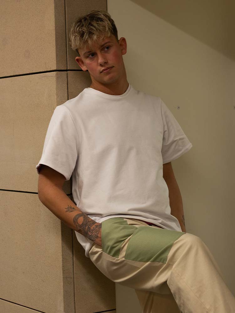 A young man in a white t-shirt and khaki pants leans against a wall.