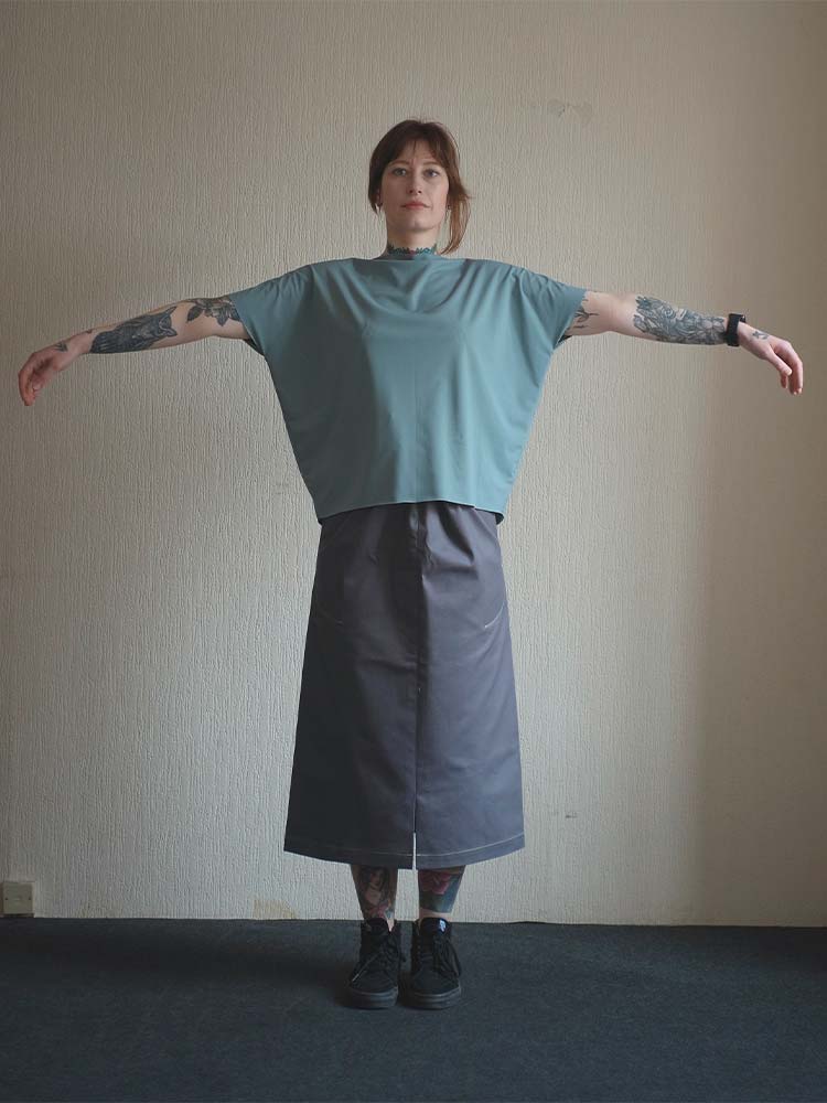 A woman models a green top and dark grey skirt in front of a wall.