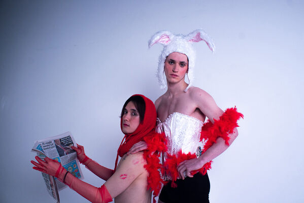 Two models, one wearing red headscarf and elbow length lace gloves reading a paper, the other stands in white corset, red feather boa and woolen rabbit ears hat