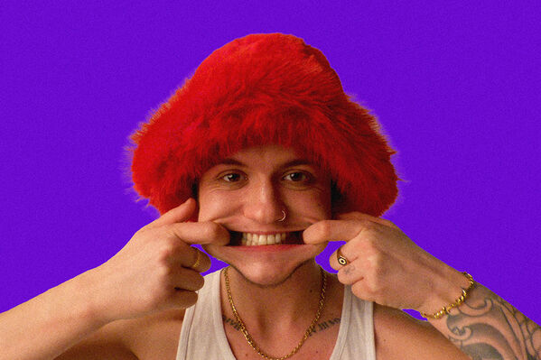 Man in white vest and red furry hat pulls the sides of his mouth with this fingers to form a wide 'grin'