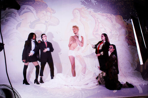 Male model wearing white trunks with long lace scarf around his neck, attended by four dark suited young people, strikes a pose in front of a projection of 'The Birth of Venus'