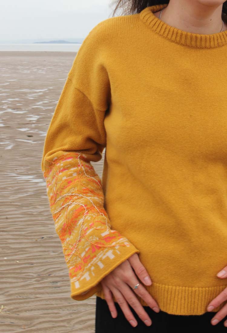 Woman in yellow sweater with embroidered sleeve and black jeans standing on beach.