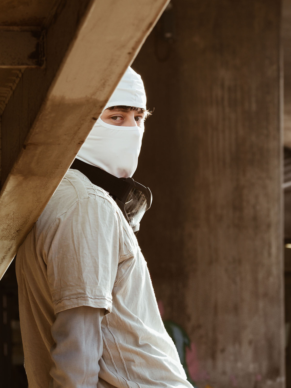 Man in white balaclava with protective goggles hanging around his neck leans against a roof beam