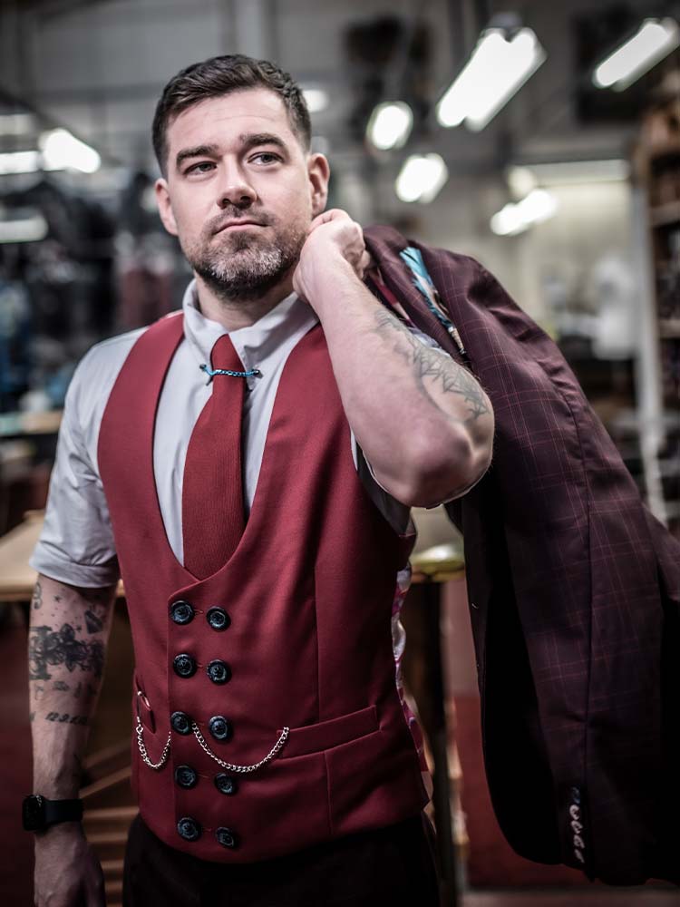 A man adorned with tattoos wearing a red waistcoat throws his jacket over his shoulder.