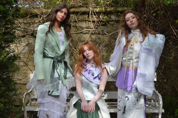 Three women model bridal wear made from hand dyed, second hand wedding dresses and silk fabrics by of a white garden bench.