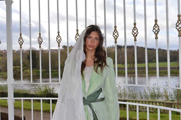 A woman models a bridal outfit made from hand dyed, second hand wedding dresses and silk fabrics featuring a full length white dress with a pale green jacket with dark green belt and long veil draped over one shoulder.