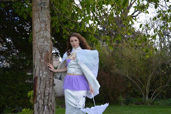 A woman models a bridal outfit made from hand dyed, second hand wedding dresses and silk fabrics featuring a purple and white skirt and top covered by a pale blue box shouldered jacket and lilac parasol
