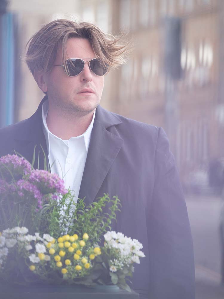 A man in sunglasses and a black jacket standing behind colorful flowers.