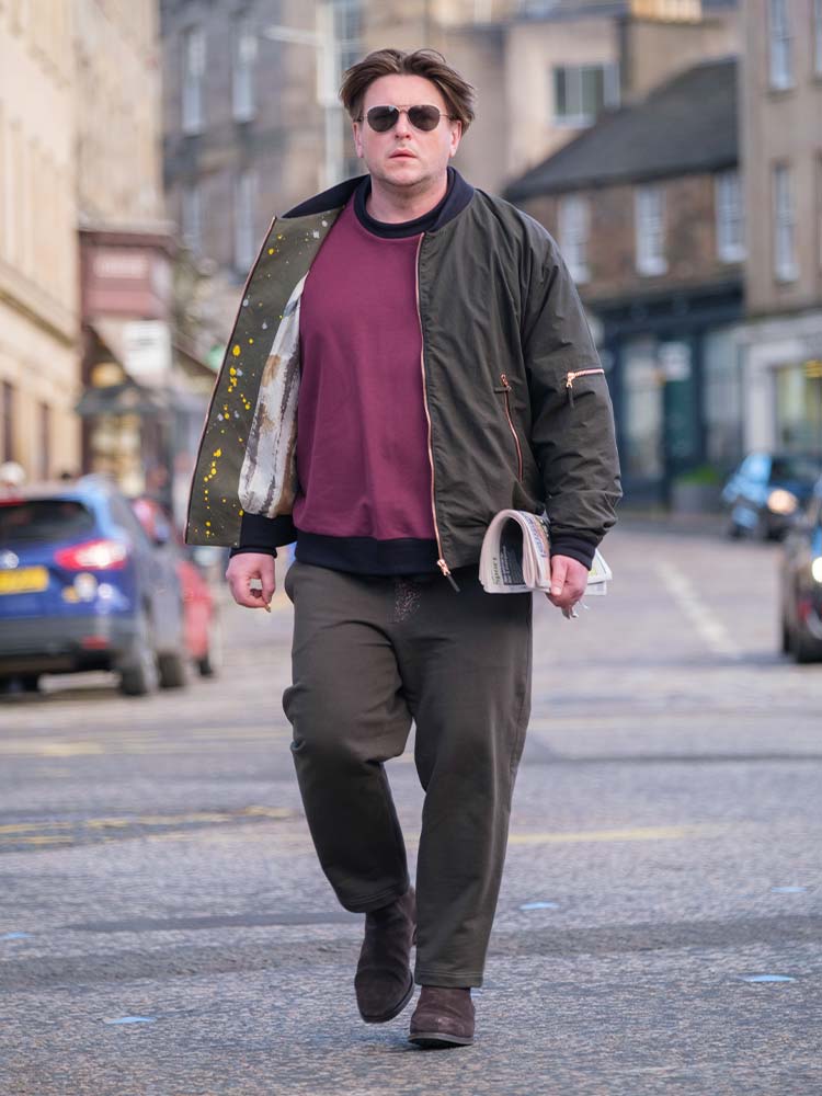 A man in a maroon sweater under a bomber jacket and brown trousers walking down the street.