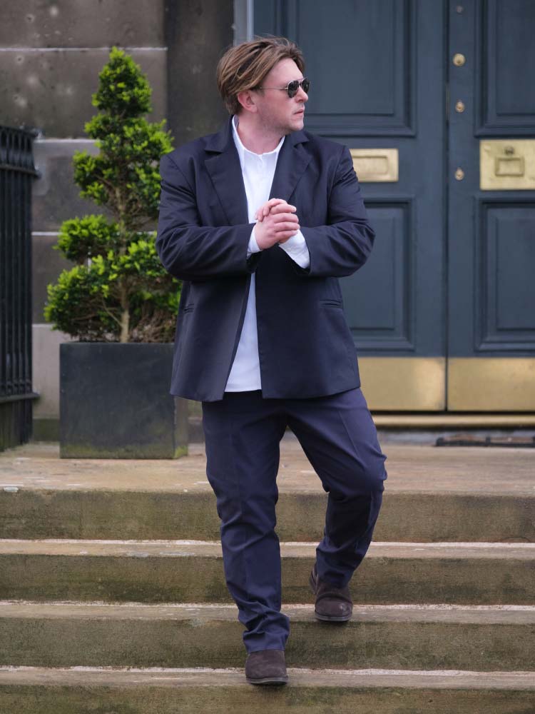 A stylish man in a suit and sunglasses stands confidently on a set of steps.