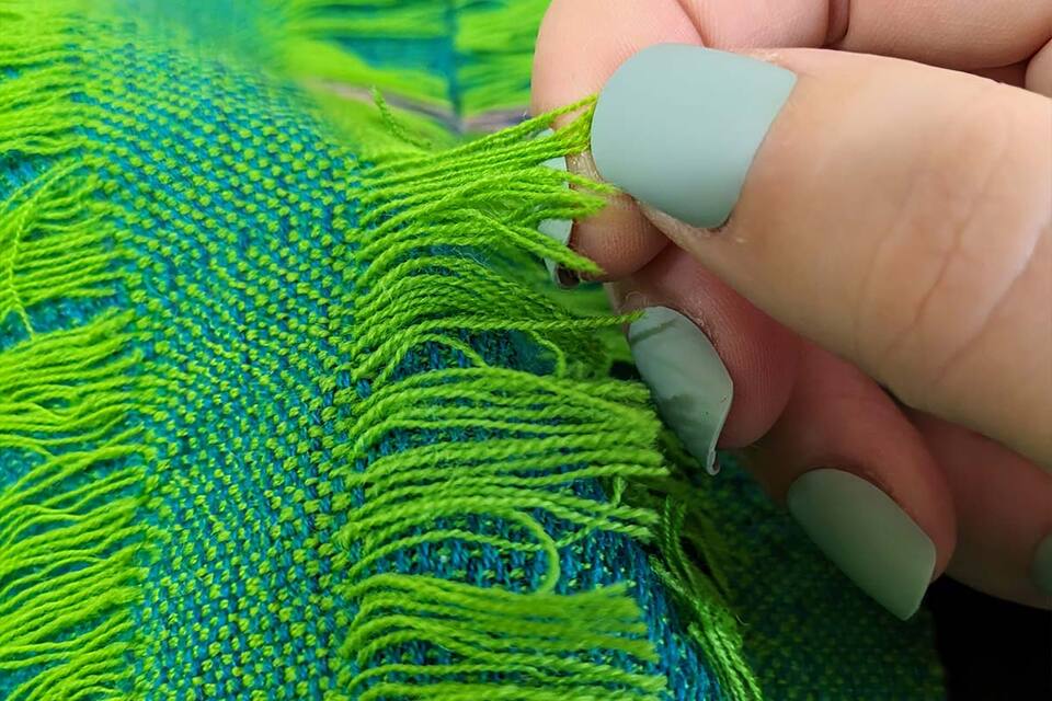 Close up of a woman teasing the fibres of a vibrant green and blue scarf using her finger and thumb.