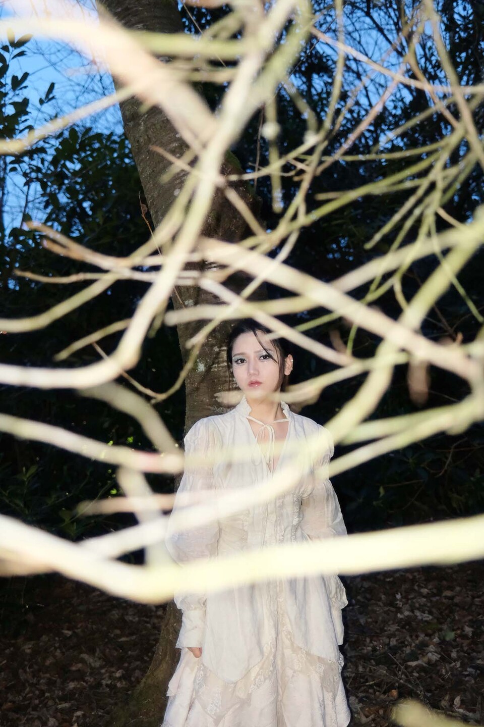Young woman in long white dress under a tree is framed by branches that criss-cross her